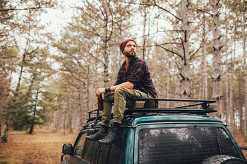 Millennial man hipster with beard sitting on car roof drinking beer in a pine forest.