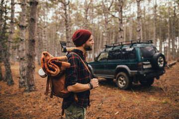 Millennial man hipster with beard wearing backpack, holding axe and firewood in forest with car on background.