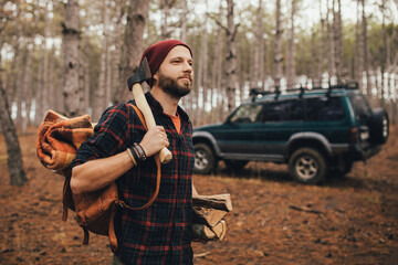 Millennial man hipster with beard wearing backpack, holding axe and firewood in forest with car on background.