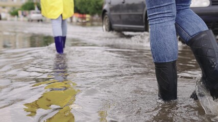 girls in raincoats and rubber boots walk along road flooded with torrential rains, their feet walk...
