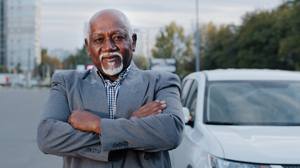 Old stately handsome african american man closeup stands near white car elderly person folded arms across chest serious face businessman ethnic male gray beard in city outdoors retiree posing thinks - Powered by Adobe