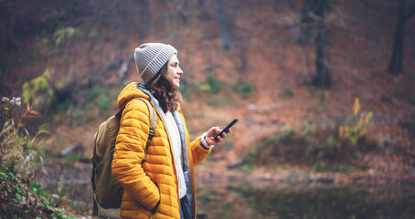Young woman traveler in yellow jacket and hat with backpack with mobile phone smartphone in hands