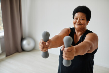 Waist up portrait of active senior woman lifting dumbbells while working out indoors and smiling at...