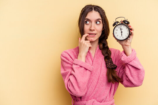 Young caucasian woman wearing a bathrobe holding a alarm clock isolated on yellow background relaxed thinking about something looking at a copy space.