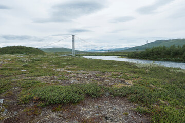 Fototapeta na wymiar Northern landscape in Swedish Lapland with pedestrian suspension bridge over Vuojatadno river, birch tree forest and green mountains. Summer overcast day, moody sky
