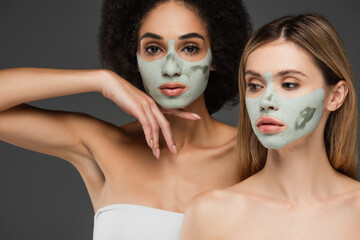 african american woman in clay mask looking at camera near friend isolated on grey