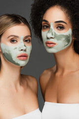 young multiethnic women looking at camera while pampering faces with clay masks isolated on grey