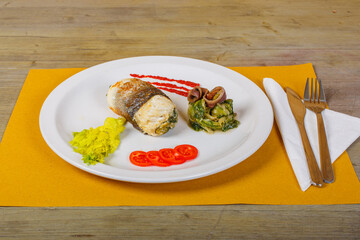 Involtino di pesce spada ripieno di verdure e mozzarella con contorno di verdure e pomodori e un calice di vino bianco sullo sfondo