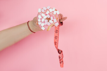 Close-up of woman's hand holding donut and measuring tape on pink background.