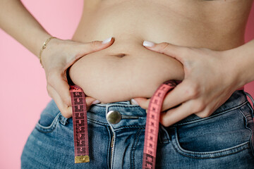 Measuring tape around waist, dieting concept, pink background, jeans, fat fold in hand