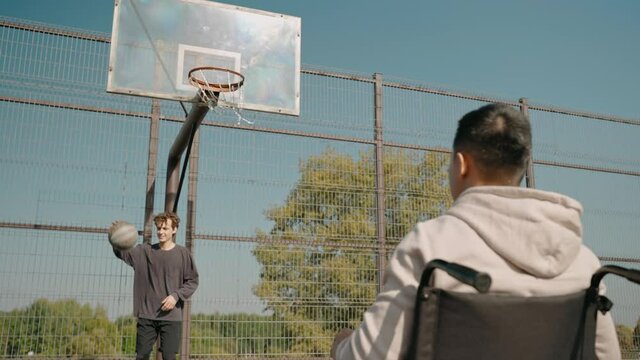 Disabled man in wheelchair playing basketball outside with his friend. Man scoring a basketball sitting in a wheelchair. High quality 4k footage