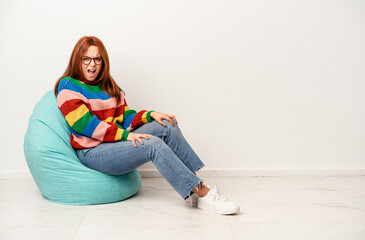 Fototapeta premium Young caucasian woman sitting on puff isolated on white background screaming very angry and aggressive.