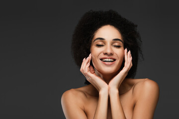 happy african american woman with closed eyes touching perfect skin on face isolated on grey