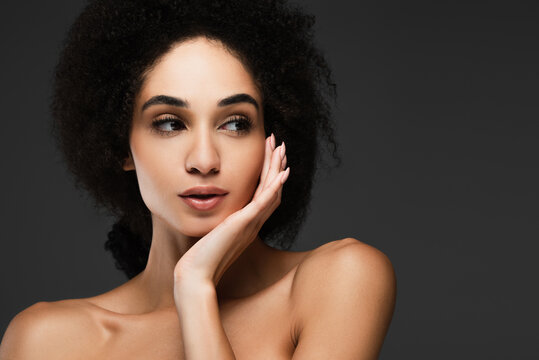 pretty african american woman with bare shoulder looking away while touching clean face isolated on grey