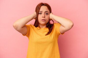 Young caucasian woman isolated on pink background covering ears with hands trying not to hear too loud sound.