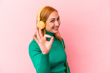 Young caucasian woman listening to music isolated on pink background cheerful and confident showing...
