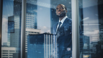 Thoughtful African-American Businessman in a Perfect Tailored Suit Standing in His Office Looking out of the Window on Big City. Successful Investment Manager Planning Strategy. Outside Shot