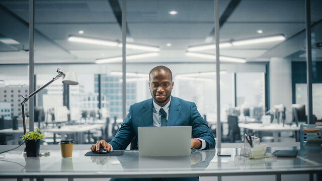 Successful Black Businessman in Tailored Suit Working on Laptop Computer on Top Floor Office Overlooking Big City. Professional CEO Managing Environmental, Social and Corporate Governance