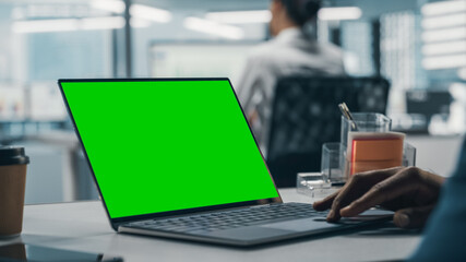 Fototapeta premium Successful Black Businessman Sitting at Desk Working on Green Screen Laptop in Office. African American Businessperson using Touch Pad on Chroma Key Computer. Shot with Focus on Hand