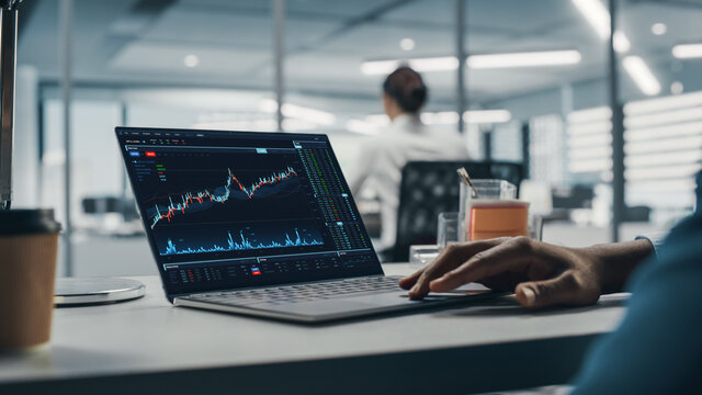 Successful Black Businessman Sitting at Desk Working on Laptop Computer in Big City Office. Hard Working Top Manager Doing Research for e-Commerce Project with Graps, Charts and Tables. Over Shoulder