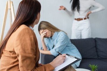 Fototapeta premium psychologist writing on clipboard near depressed lesbian woman and her african american girlfriend