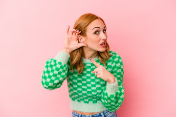 Young caucasian woman isolated on pink background trying to listening a gossip.