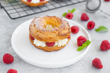 Choux pastries. Choux rings with cream of cream cheese or cottage cheese and fresh raspberries, dusted with powdered sugar on a white plate on a gray concrete background.