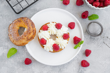 Choux pastries. Choux rings with cream of cream cheese or cottage cheese and fresh raspberries, dusted with powdered sugar on a white plate on a gray concrete background.