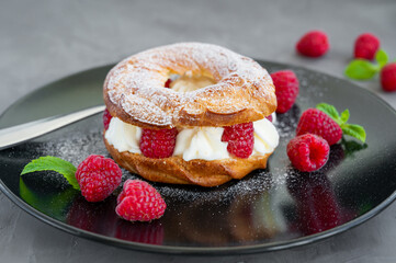 Choux pastries. Choux rings with cream of cream cheese or cottage cheese and fresh raspberries, dusted with powdered sugar on a black plate on a gray concrete background.