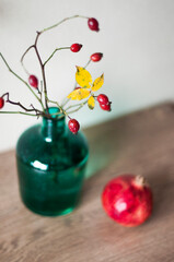 Turquoise glass vase with branches and rose hips berries, yellow leaves and ripe pomegranate on a wooden surface against a background of beige wall top view.