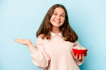 Little caucasian girl holding a bowl of cereals isolated on blue background showing a copy space on a palm and holding another hand on waist.