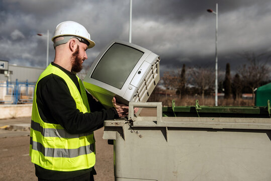 Trabajador del centro de reciclaje arrojando un viejo televisor de tubo en un contenedor de basura gris. Hombre que lleva un chaleco reflectante y casco protector. Reciclaje de aparatos electr&oacute;nicos