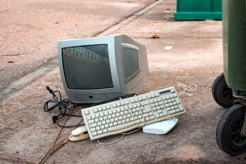 Televisión de tubo vieja y otros aparatos electrónicos, como un teclado antiguo en el suelo de la calle junto a un contenedor de basura verde. Concepto de reciclaje de tecnología - obrazy, fototapety, plakaty