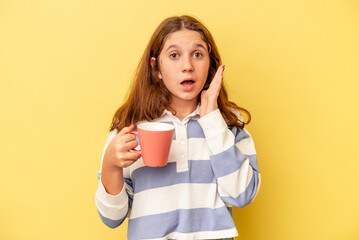 Little caucasian girl holding a pink mug isolated on yellow background surprised and shocked.