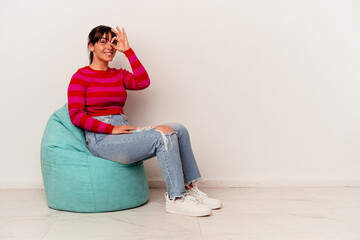 Young Argentinian woman sitting on a puff isolated on white background excited keeping ok gesture on eye.