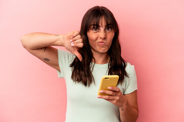 Young Argentinian woman holding a mobile phone isolated on pink background showing a dislike gesture, thumbs down. Disagreement concept.