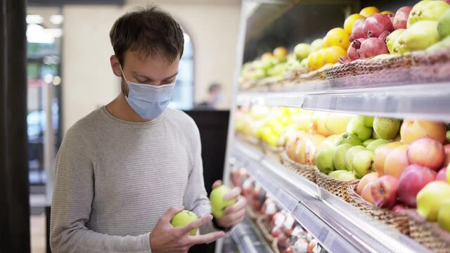 Man in mask takes a fresh apple from the food shelf. Shopper choosing apple at grocery store
