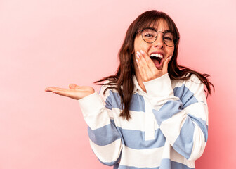 Young Argentinian woman isolated on pink background holds copy space on a palm, keep hand over cheek. Amazed and delighted. - obrazy, fototapety, plakaty