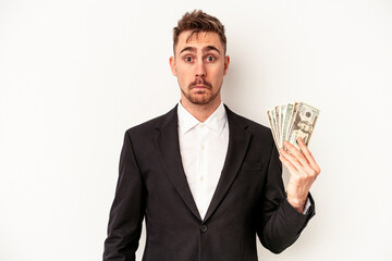 Young caucasian business man holding bank notes isolated on white background shrugs shoulders and open eyes confused.