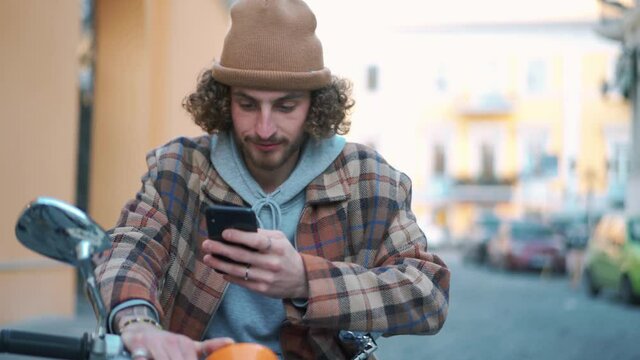 Cheerful curly-haired man texting by phone while sitting on moped outdoors