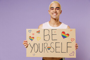Young latin gay man with make up wearing bright pink top hold card sign with be yourself title text isolated on plain pastel purple background studio portrait. People lifestyle fashion lgbtq concept.