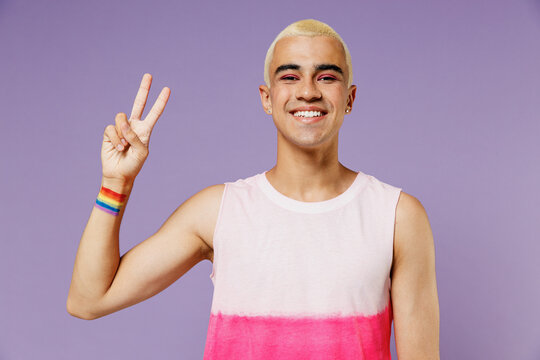 Young smiling friendly latin gay man 20s with make up wearing bright pink top showing victory sign isolated on plain pastel purple background studio portrait. People lifestyle fashion lgbtq concept.