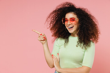 Fototapeta premium Promoter young curly latin woman 20s years old wear mint t-shirt sunglasses point index finger away on workspace area copy space mock up isolated on plain pastel light pink background studio portrait