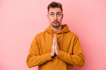 Young caucasian man isolated on pink background holding hands in pray near mouth, feels confident.