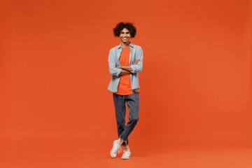 Full body young smiling black student man 50s wearing blue shirt t-shirt looking camera hold hands crossed folded isolated on plain orange color background studio portrait. People lifestyle concept
