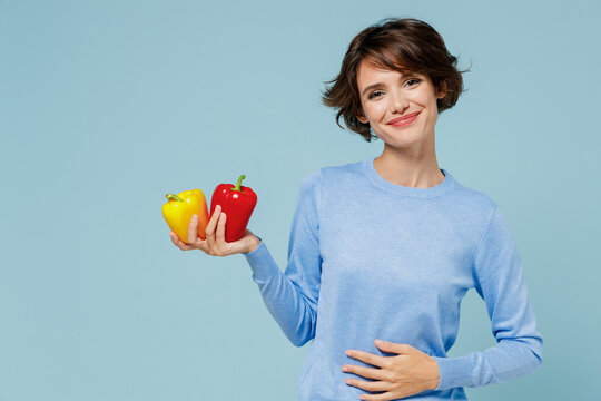 Young Satisfied Happy Caucasian Woman 20s In Casual Sweater Look Camera Hold Two Yellow Red Bell Peppers Isolated On Plain Pastel Light Blue Background Studio Portrait. People Lifestyle Food Concept.