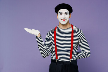 Smiling vivid young mime man with white face mask wears striped shirt beret pointing aside on empty...