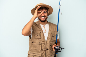 Young mixed race fisherman holding a rod isolated on blue background excited keeping ok gesture on eye.