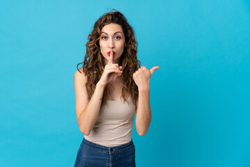 Young caucasian woman isolated on blue background pointing to the side and doing silence gesture