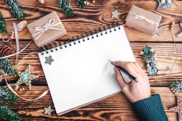 A female hand writes New Year's plans into a notebook on the background of a wooden table with gifts and New Year's decor.